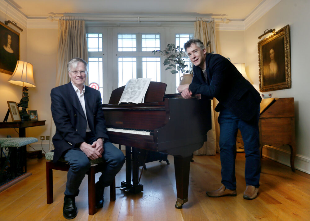 Two men in a cozy living room pose by a grand piano: one seated in a suit, the other leaning on the piano with sheet music open between them.