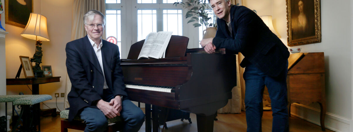 Two men in a cozy living room pose by a grand piano: one seated in a suit, the other leaning on the piano with sheet music open between them.