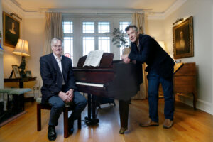 Two men in a cozy living room pose by a grand piano: one seated in a suit, the other leaning on the piano with sheet music open between them.