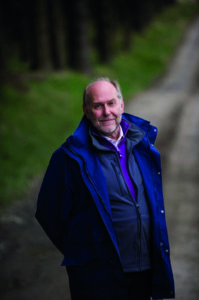 Older man in a blue jacket standing outdoors on a winding path, greenery in the background and soft light on his face.