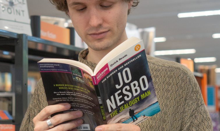 Young man with curly hair reads a Jo Nesbo novel, 'The Jealous Man,' in a bookstore.
