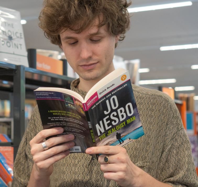 Young man with curly hair reads a Jo Nesbo novel, 'The Jealous Man,' in a bookstore.