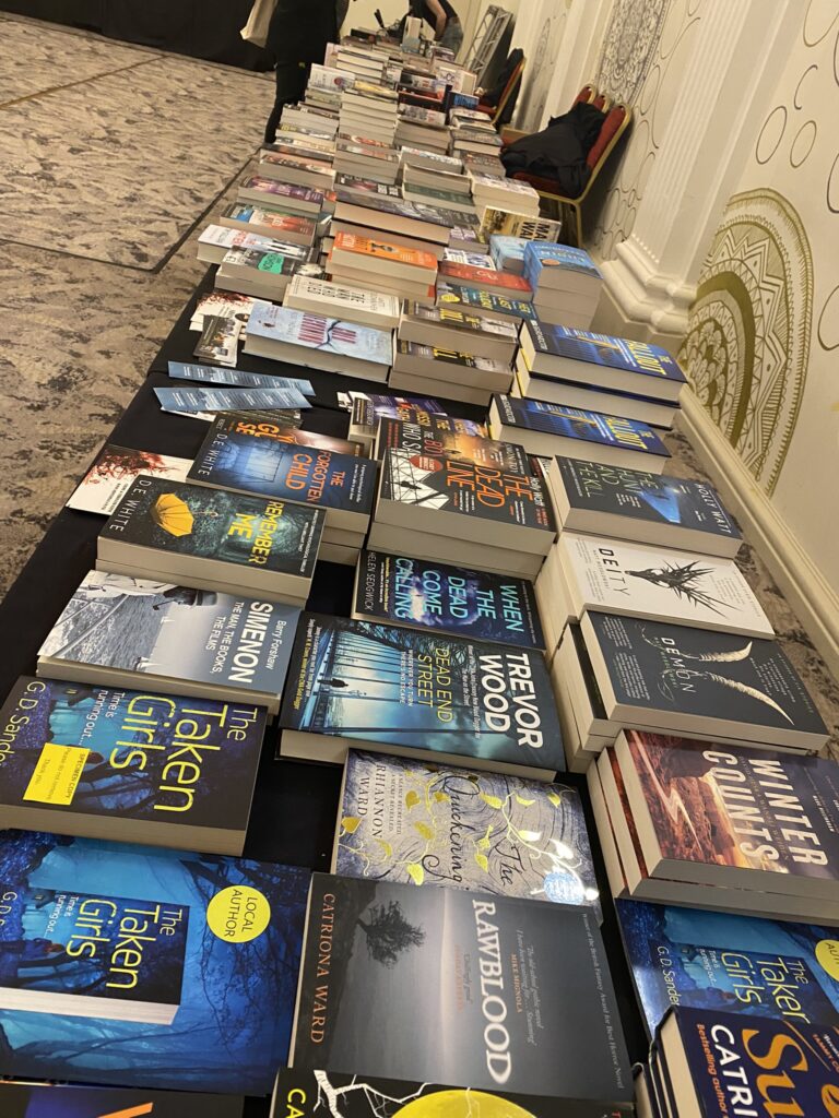 Table lined with stacked hardcover and paperback books at a book fair, arranged in neat rows along a black cloth-covered surface in a patterned room. National Crime Reading Month
